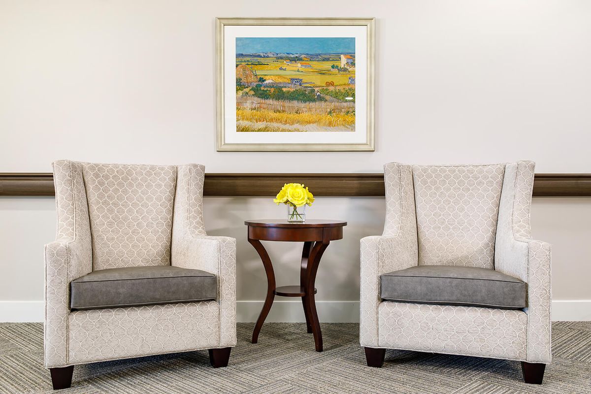 A cozy interior setting featuring two upholstered chairs, a small round table with a yellow flower arrangement, and a framed painting on the wall.