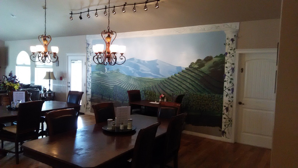 A dining room with a vineyard mural covering one wall, a chandelier hanging from the ceiling, and a wooden table surrounded by chairs.