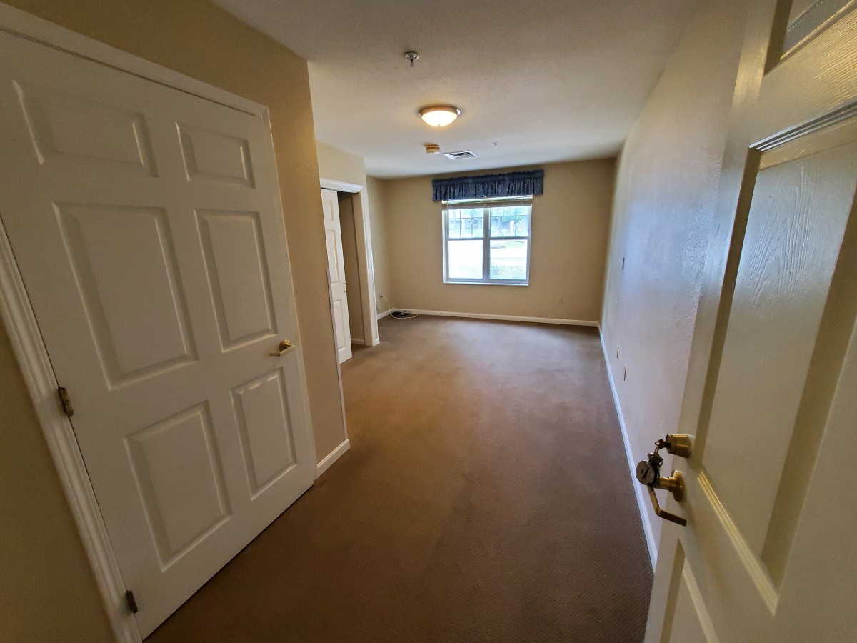 A vacant room with beige walls and carpet, featuring a window and a closed door.