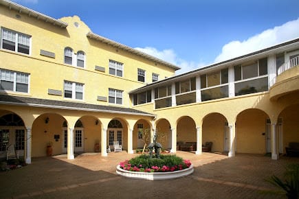 A sunny courtyard with a central fountain surrounded by flowers, framed by a yellow building with arches.