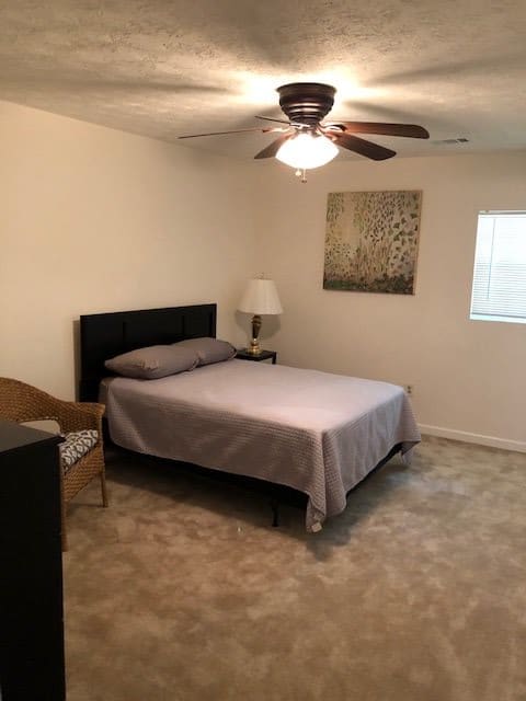 A simple bedroom with a bed, lamp, chair, and ceiling fan.