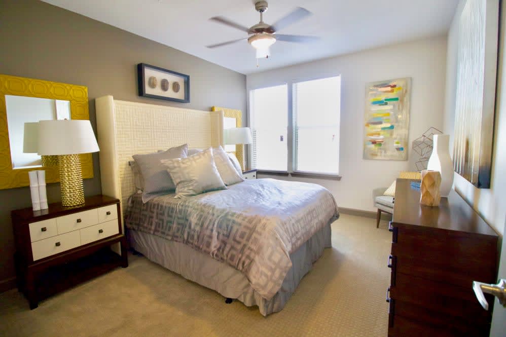 A cozy and well-decorated bedroom featuring a bed with patterned bedding, bedside tables, lamps, artwork on the walls, and a ceiling fan.
