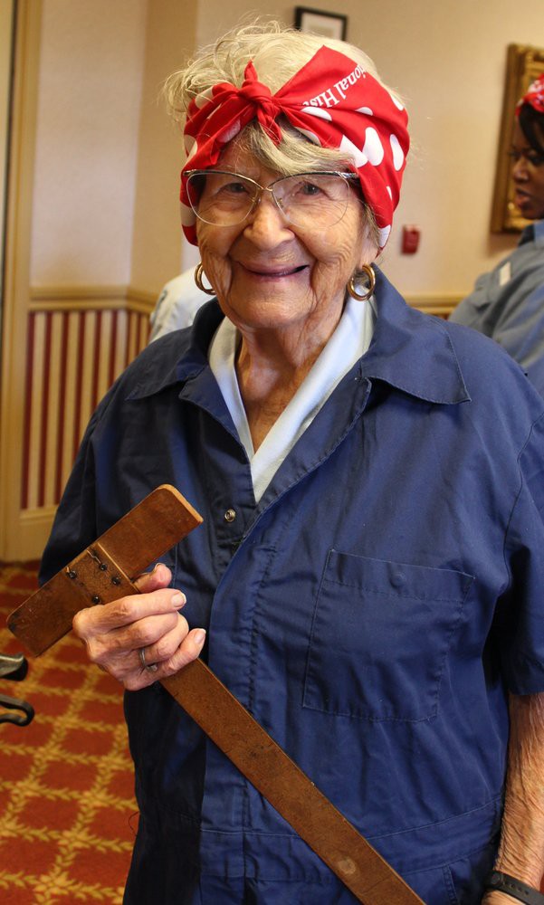 Elderly woman with a red bandana and glasses, smiling while holding a wooden item.