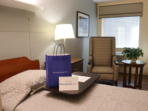 A cozy hotel room featuring a bed with a decorative lamp, a chair for relaxation, and a gift bag on the table.