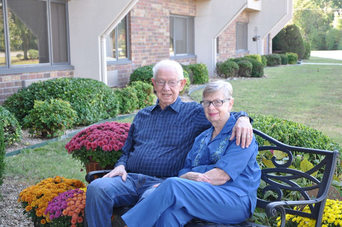 An elderly couple sitting together on a bench in a garden setting.