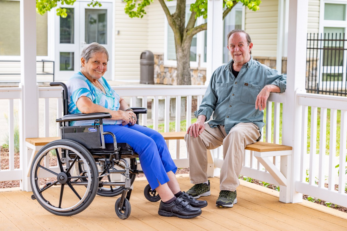 An elderly woman in a wheelchair sitting next to a man on a bench, enjoying time outdoors.