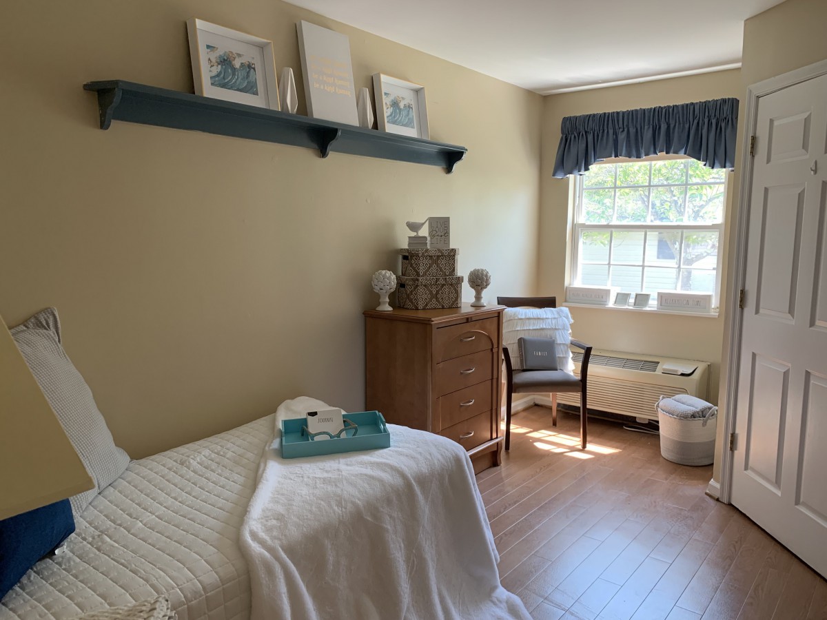 A cozy bedroom with natural light, featuring a bed, wooden furniture, and simple decor.