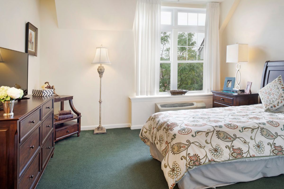 A cozy bedroom with a large window, featuring a bed with patterned bedding, a standing lamp, and wooden furniture.