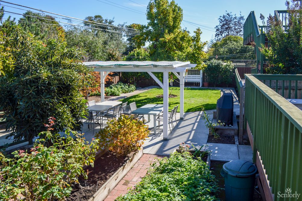 A tranquil backyard features a white pergola with outdoor seating, surrounded by lush greenery and garden on a sunny day.
