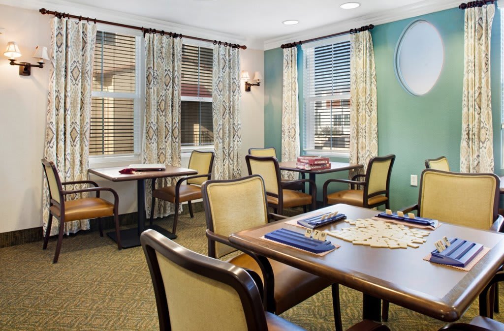 A cozy game room with tables and chairs, featuring windows with curtains and board games on the tables.