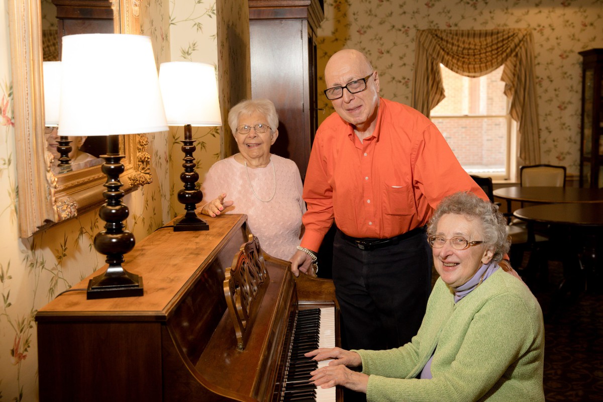A group of seniors enjoying time together around a piano in a cozy living room.