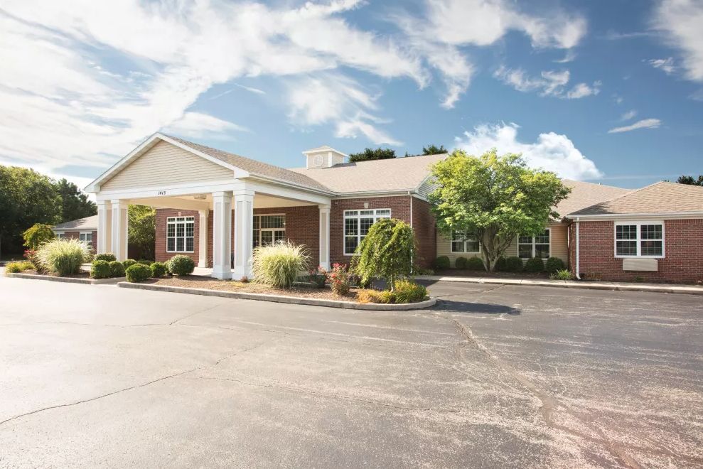 A single-story brick building with white columns, surrounded by landscaping and a parking lot under a partly cloudy sky.