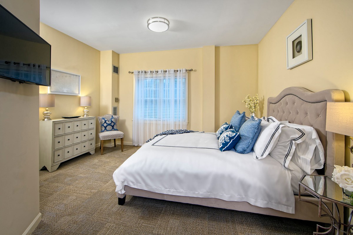 A cozy bedroom with soft lighting and neutral tones, featuring a comfortable bed, dresser, and chair.