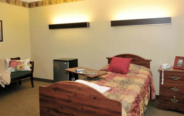 A cozy bedroom with a wooden bed, colorful bedding, wall-mounted lights, and a small fridge.