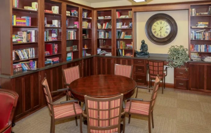 A cozy library corner with a round wooden table surrounded by chairs, bookshelves filled with books, and a large wall clock.