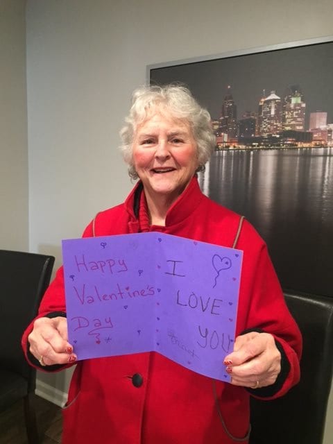 An elderly woman wearing a red coat smiles while holding a greeting card indoors.