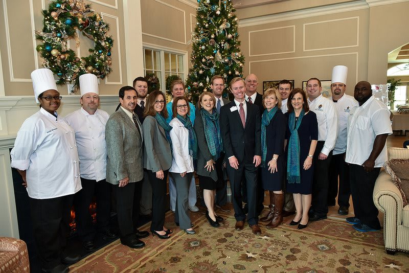 A group of people in formal attire and chefs' uniforms posing in front of a decorated Christmas tree indoors.