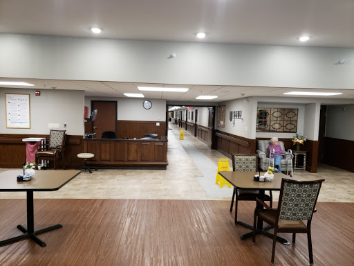 A spacious common area in a senior living facility, featuring tables, chairs, and a hallway leading to other areas.