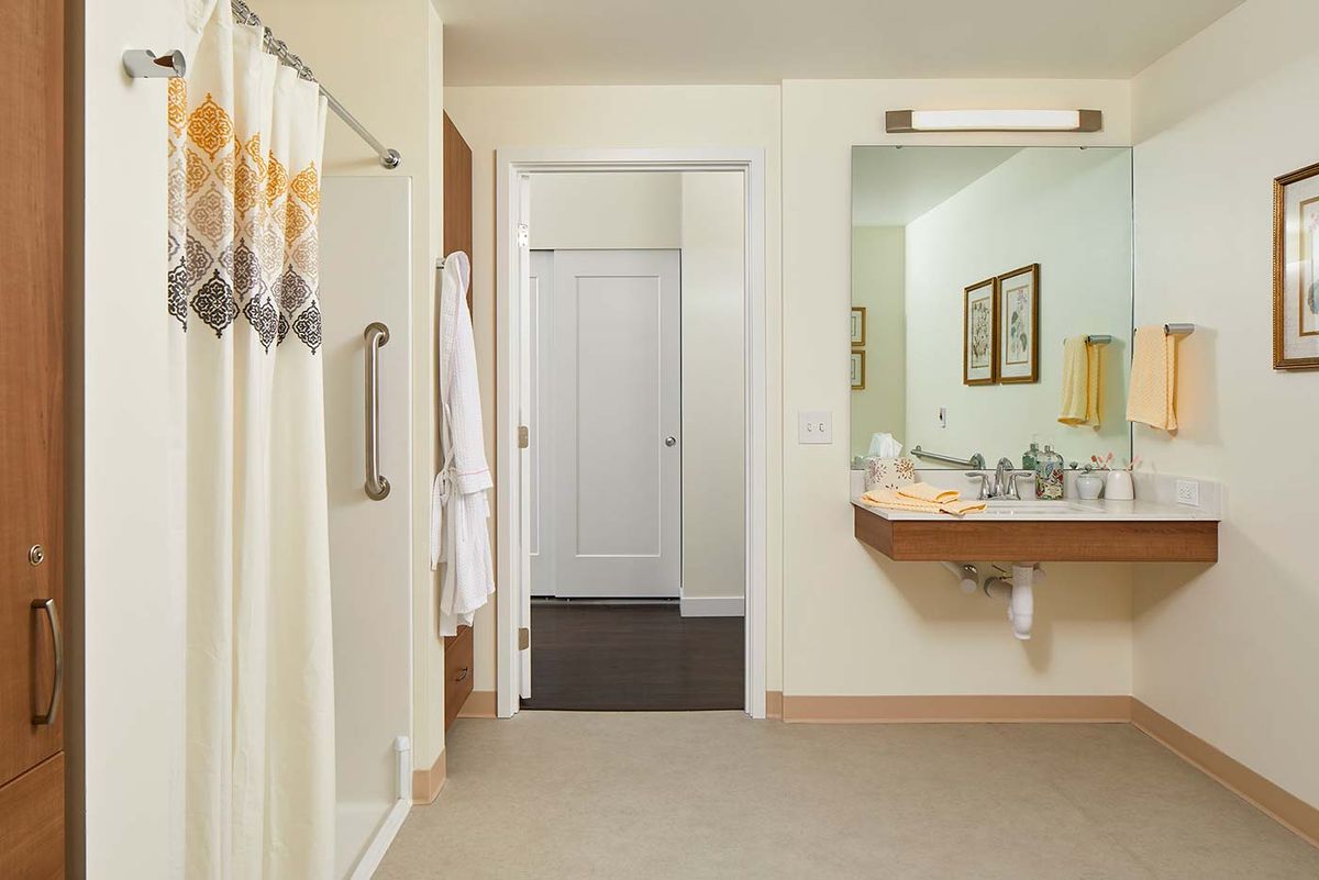 A well-lit bathroom with a shower, sink, and mirror, featuring a doorway and hanging robe.