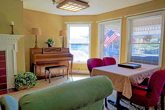 A cozy living room featuring a piano, green couch, and a table with red chairs overlooking an American flag outside the window.
