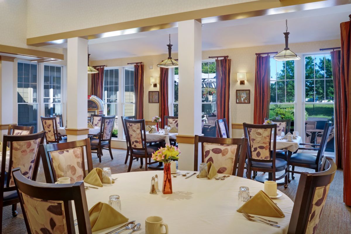 A cozy dining room with multiple tables and chairs, decorated with curtains and floral accents.