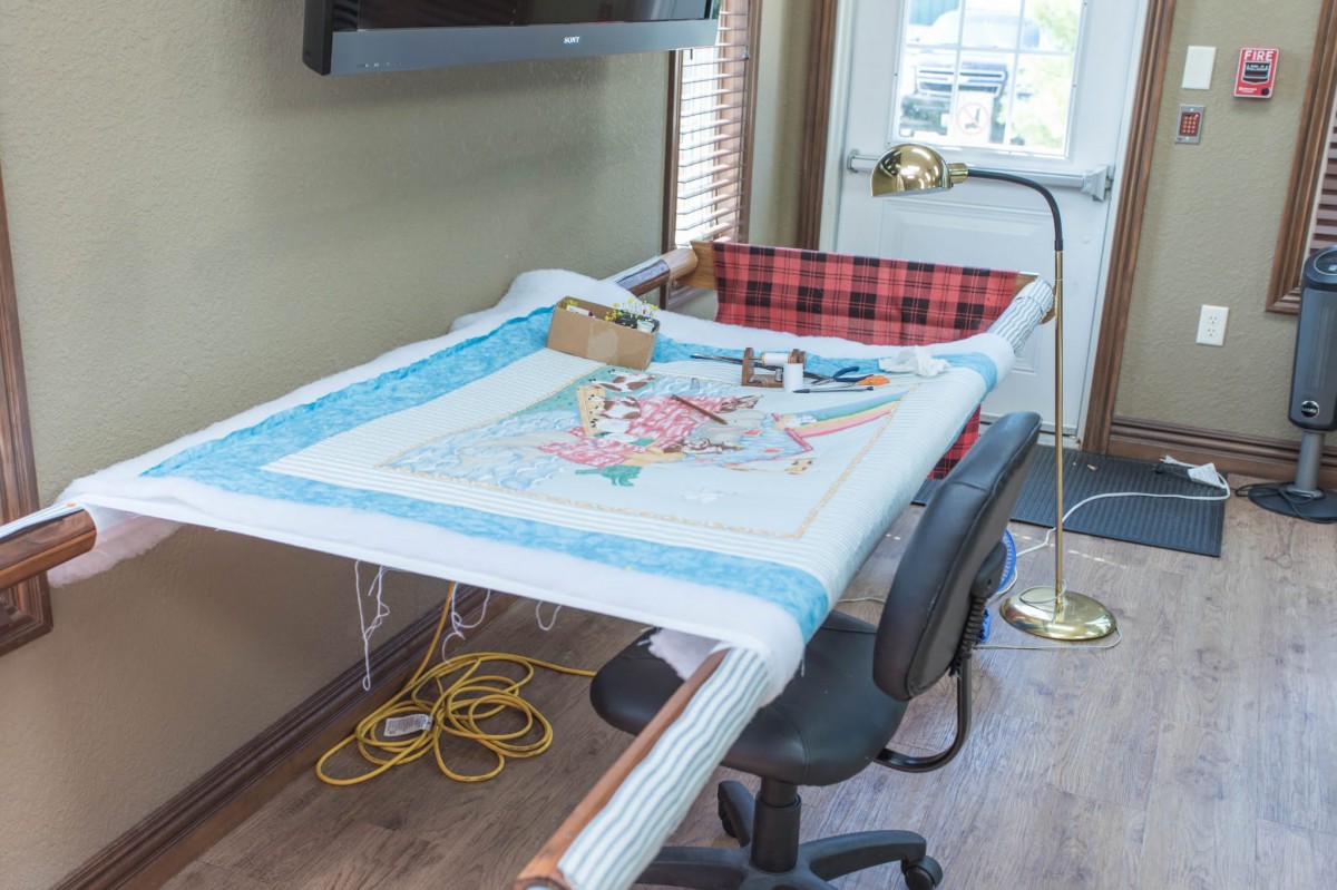A crafting workspace set up for quilting, featuring a quilt in progress on a frame, with various tools and a lamp nearby.