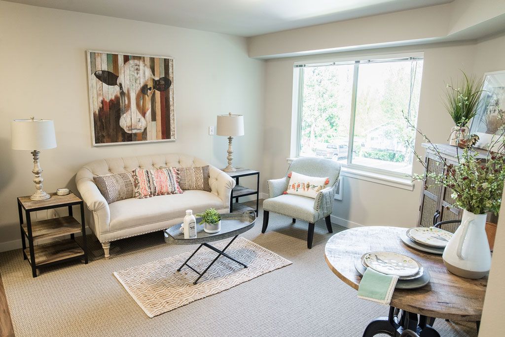 A cozy living room featuring a couch, armchair, coffee table, and a large window. The walls are adorned with artwork that adds character to the space.