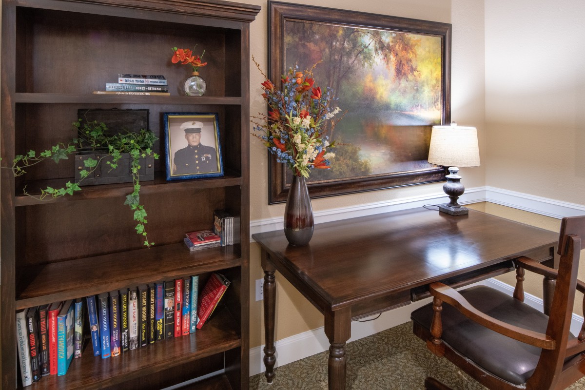 A cozy office space with a wooden desk and bookshelf, adorned with books, a vase of flowers, and a table lamp.