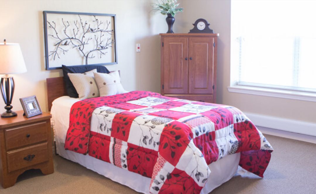 A cozy bedroom featuring a colorful quilt on the bed, a wooden cabinet, and a large window allowing natural light.