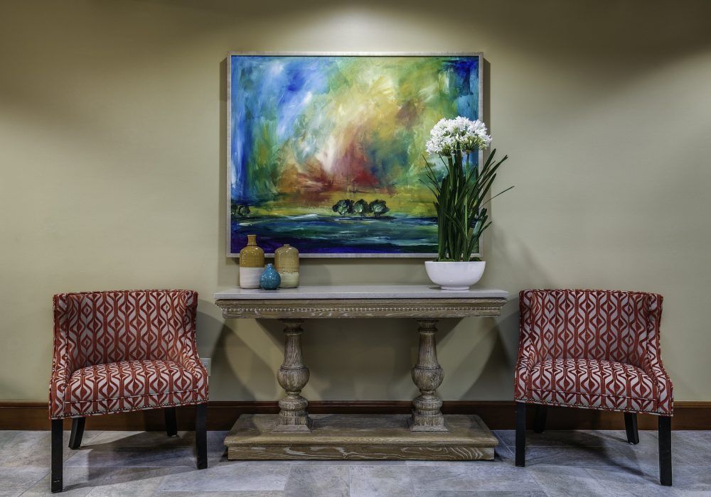 A well-decorated interior featuring a colorful painting, a table with vases and an orchid plant, flanked by two patterned chairs.