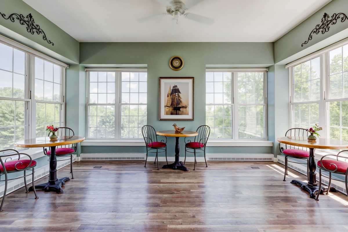 A well-lit room with large windows, featuring two small tables and chairs on a wooden floor.