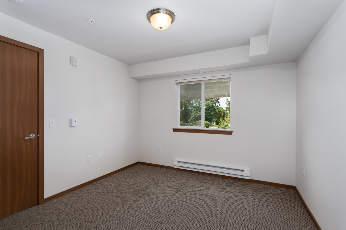 An empty room with white walls, a carpeted floor, and a window.