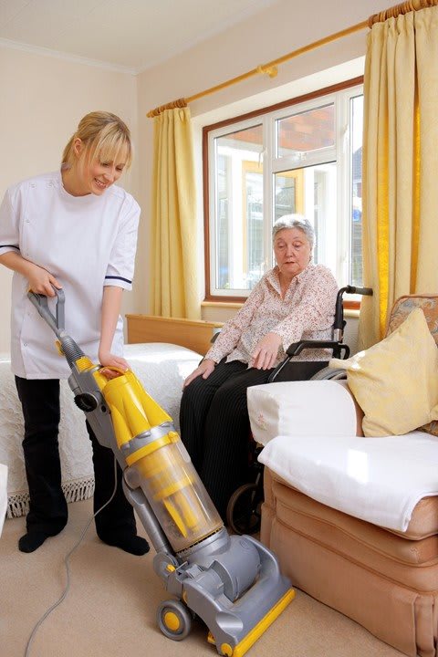 A caregiver vacuuming the room while assisting an elderly person.
