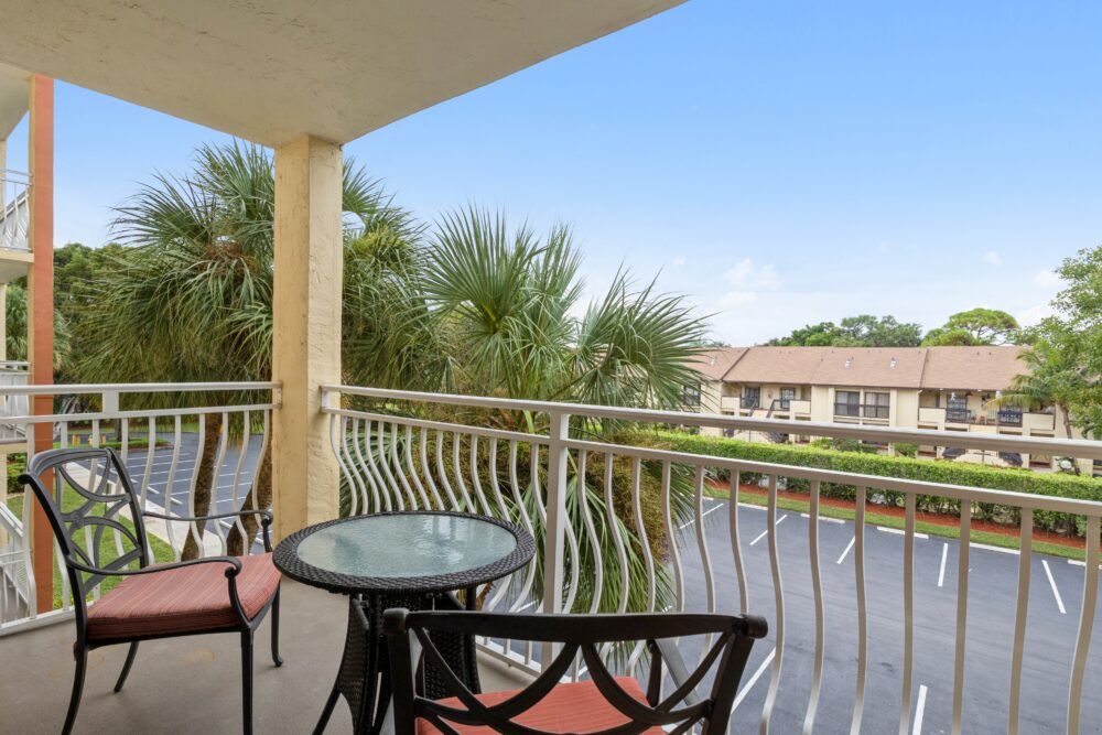 A balcony with a small table and chairs overlooking palm trees and a residential area.