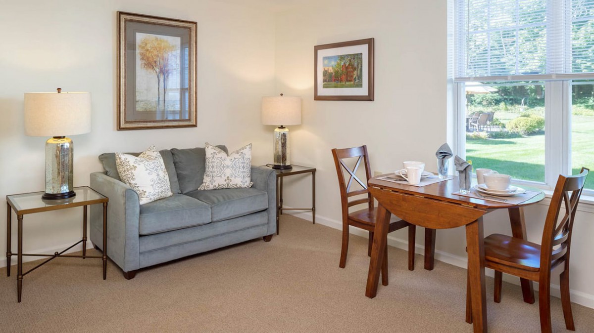 A cozy living room with a sofa, chair, and table, adorned with artwork and a lamp.