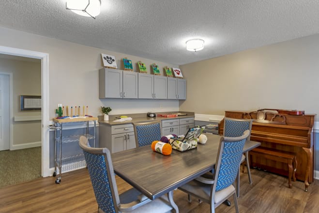 A cozy dining area with a table, chairs, and nearby art supplies, highlighted by a piano and shelving for decor.