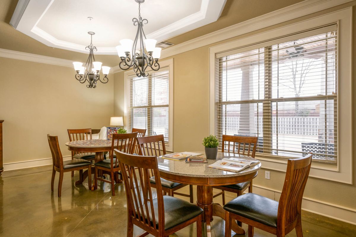 A cozy dining room featuring wooden tables and chairs under elegant chandeliers, with natural light streaming through large windows.