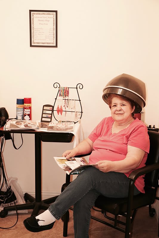 A woman sits under a hair dryer in a salon, reading a magazine.