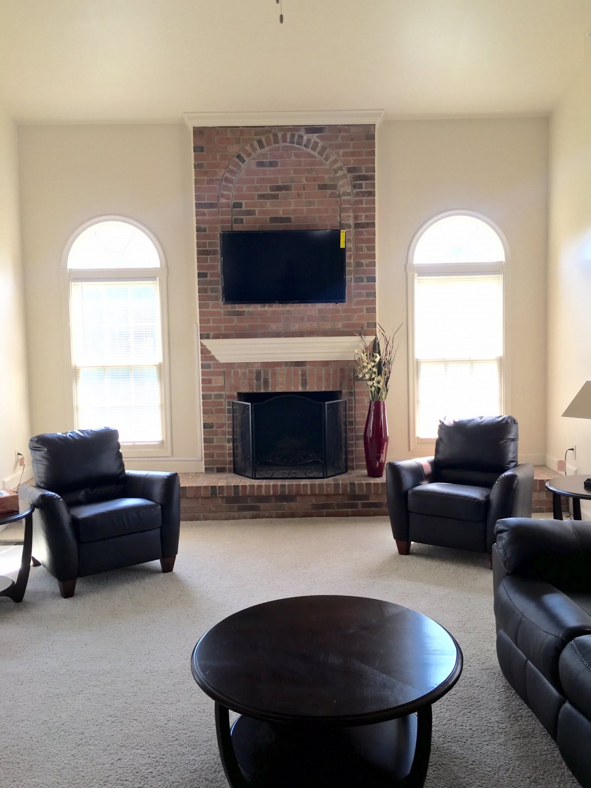 A cozy living room featuring a brick fireplace and a mounted television, flanked by two large windows and surrounded by comfortable armchairs.