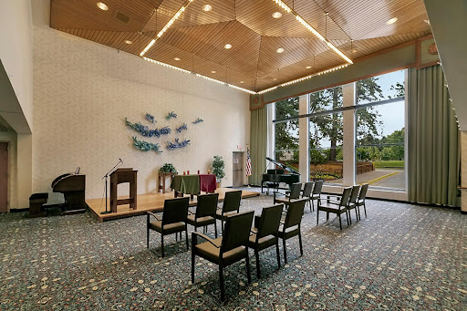 A spacious room designed for gatherings, featuring a wooden ceiling and natural lighting, with chairs arranged in front of a small stage.
