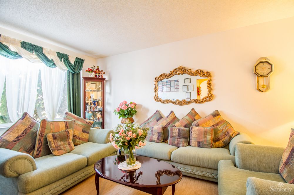 A cozy living room featuring a large sofa with patterned pillows, a decorative mirror on the wall, and a coffee table adorned with fresh flowers.