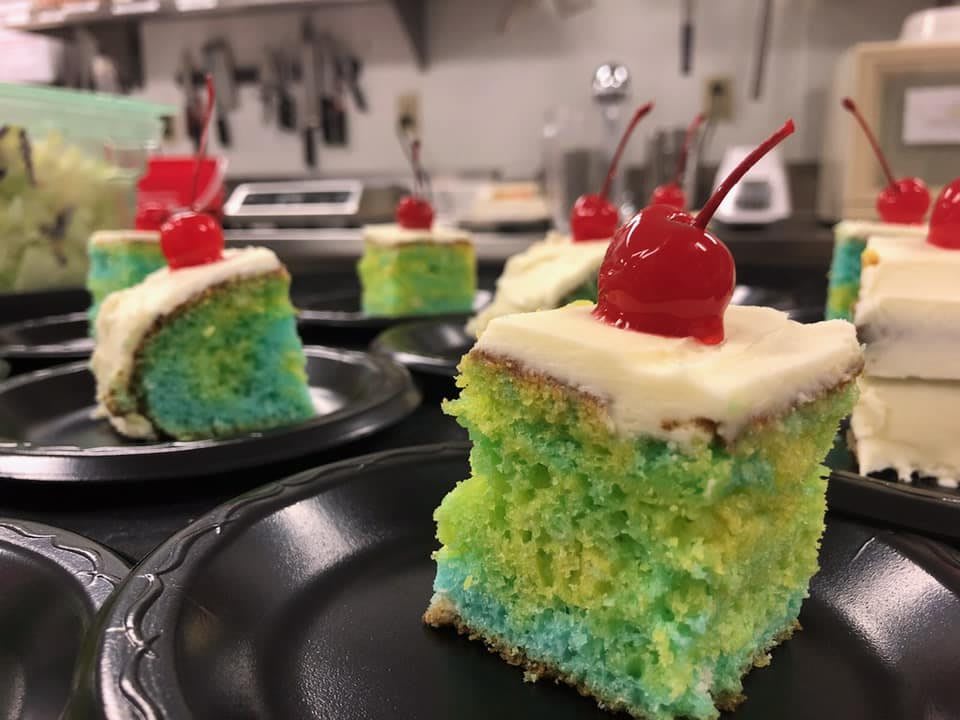 A slice of colorful cake topped with creamy frosting and a cherry.