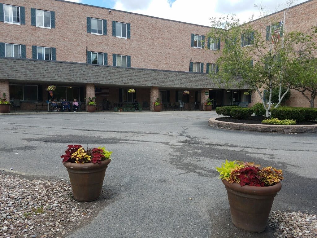 A three-story brick building with a main entrance surrounded by potted plants and a parking area.