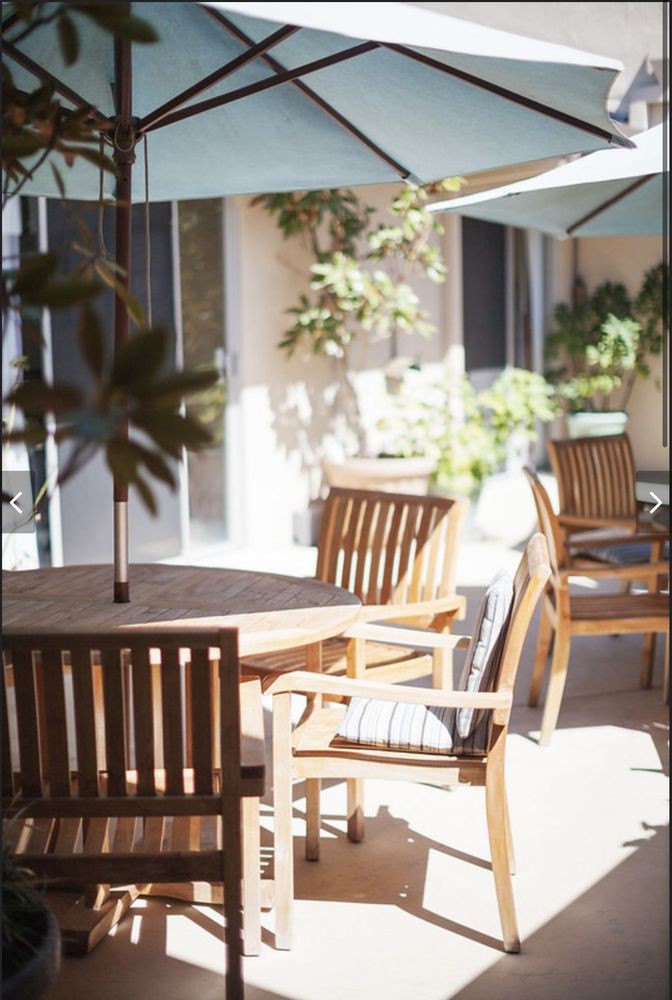 A sunlit patio featuring wooden chairs and tables under umbrellas, surrounded by greenery.