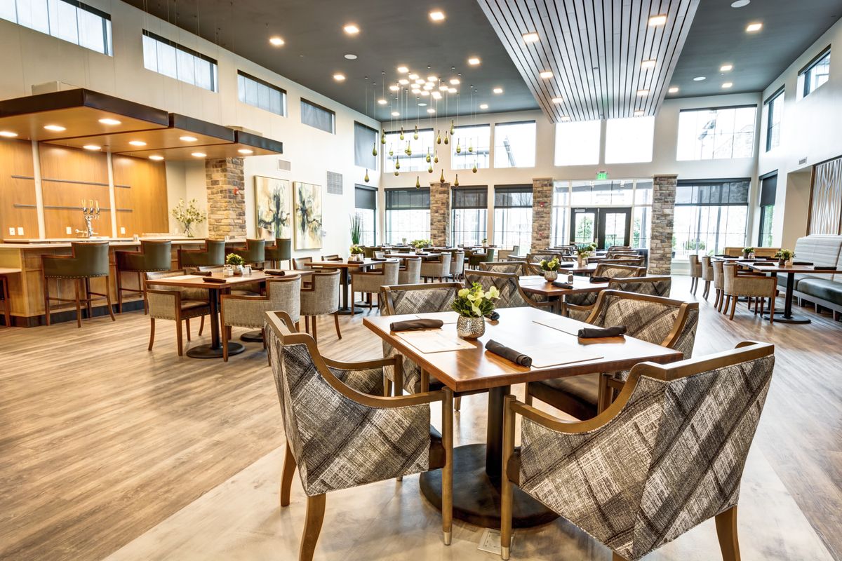 A modern restaurant interior with elegantly arranged tables and chairs, featuring a warm wooden floor and stylish lighting.