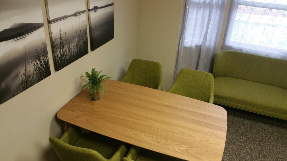 A small meeting room with a wooden table surrounded by green chairs, featuring a potted plant and artwork on the walls.