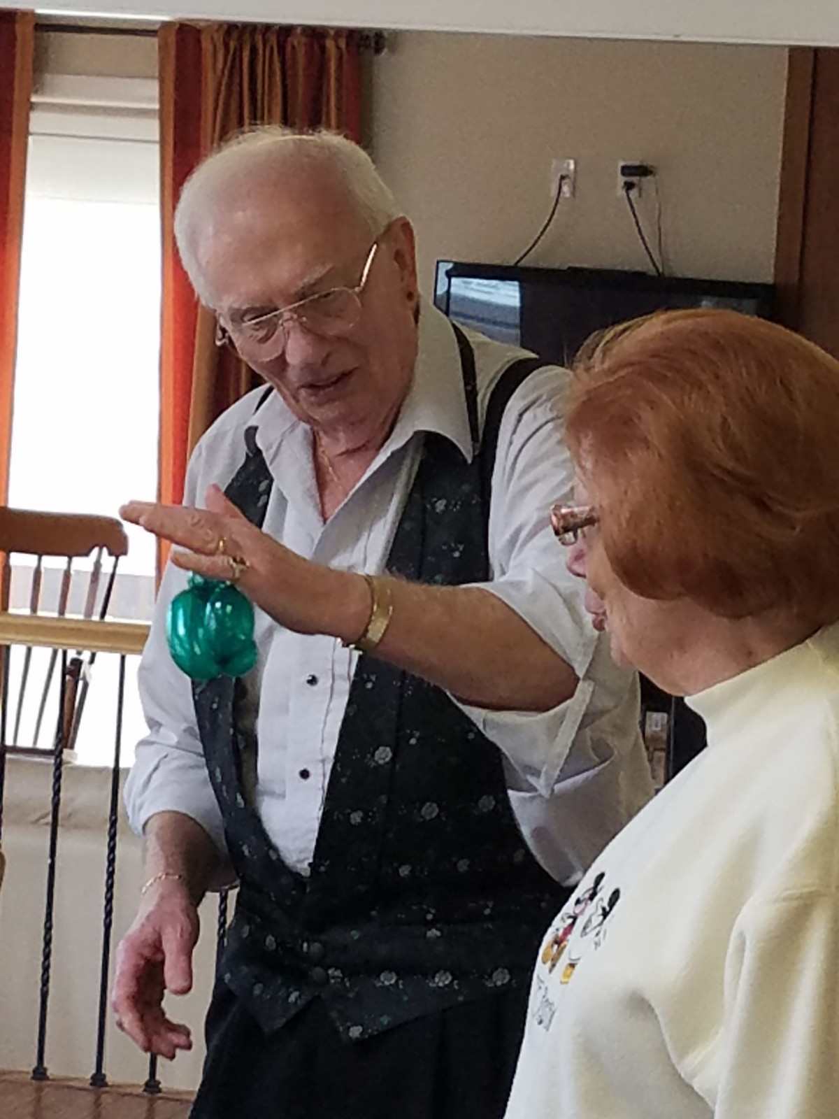 An elderly man demonstrates something to a woman in a casual indoor setting.