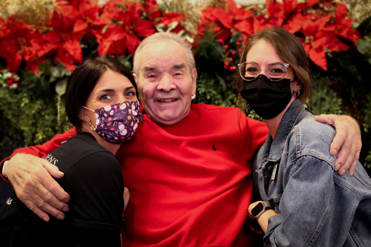 A group of three people, two of whom are wearing masks, posing happily in front of festive holiday decorations with red flowers.