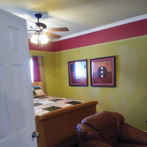 A cozy bedroom with colorful walls, featuring a bed, an armchair, wall art, and a ceiling fan.
