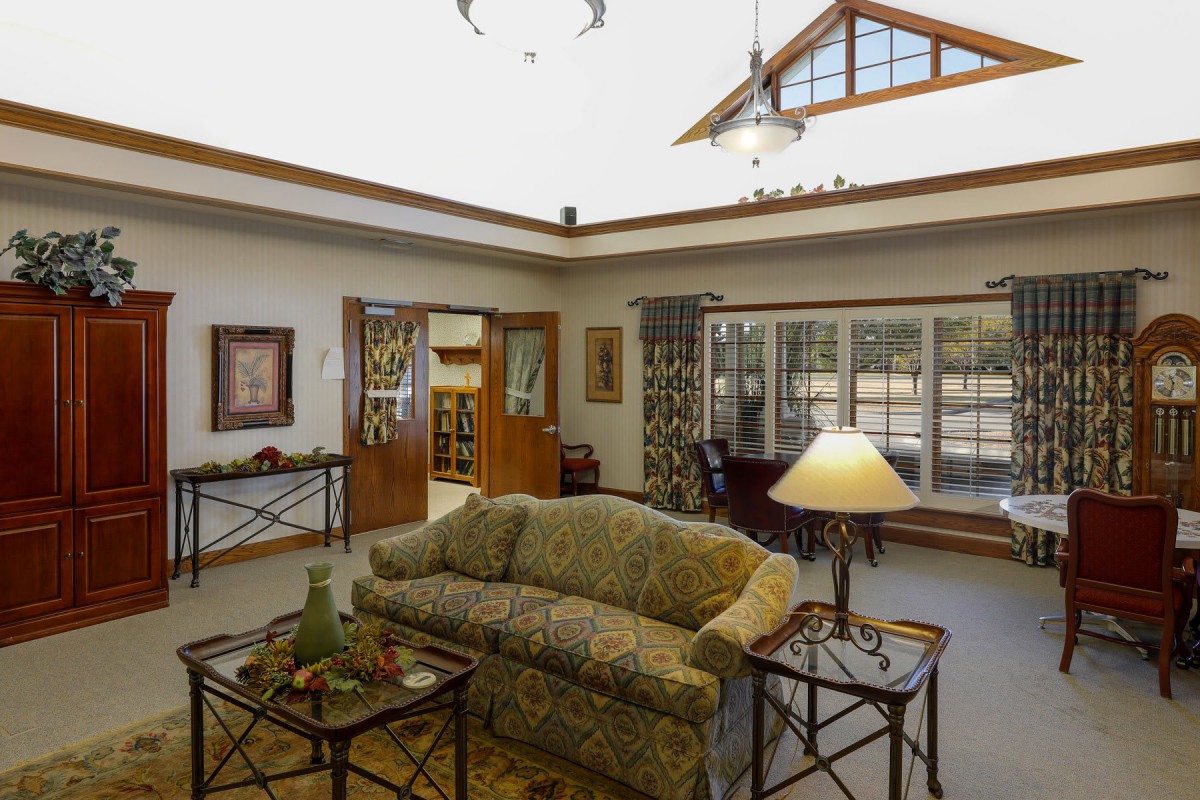 A cozy living room with a couch, tables, and a lamp, illuminated by natural light from large windows.